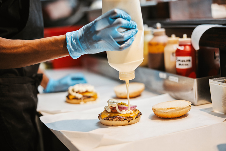 Hands, burger and sauce in fast food kitchen at counter with gloves, cooking and take away at small business. Restaurant chain, sandwich and cook with hamburger patty for lunch service with cheese